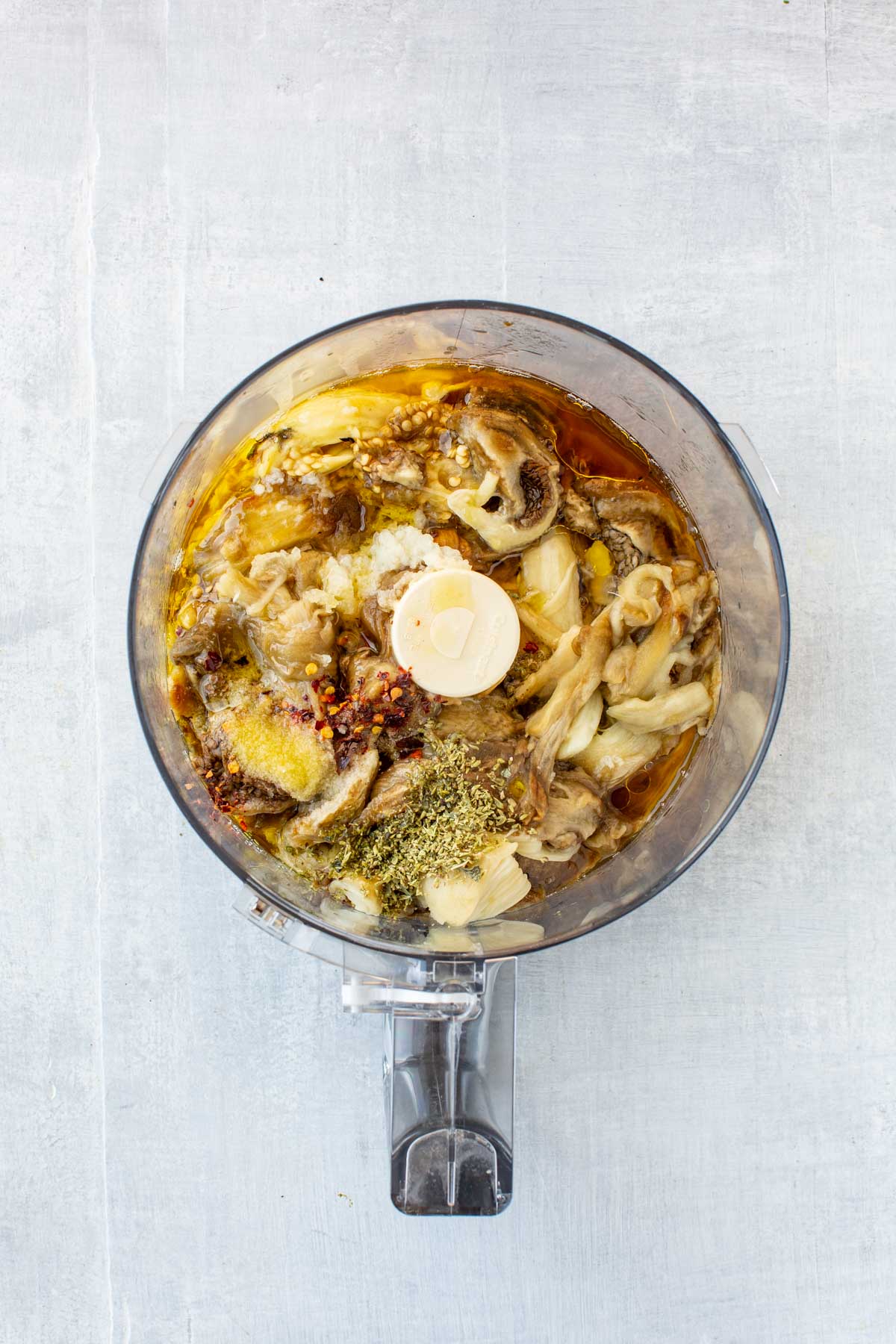 Eggplant dip ingredients in a food processor - Charred eggplant flesh with garlic cloves, oregano, red pepper flakes, and extra virgin olive oil inside the bowl of a food processor, ready to blend.