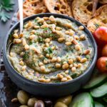 Eggplant dip with bread and vegetables - A black bowl of smoky eggplant dip with pine nuts, surrounded by cucumber slices, cherry tomatoes, olives, and crusty bread on a serving board.