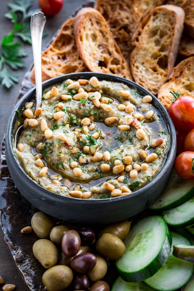 Eggplant dip with bread and vegetables – A black bowl of smoky eggplant dip with pine nuts, surrounded by cucumber slices, cherry tomatoes, olives, and crusty bread on a serving board.