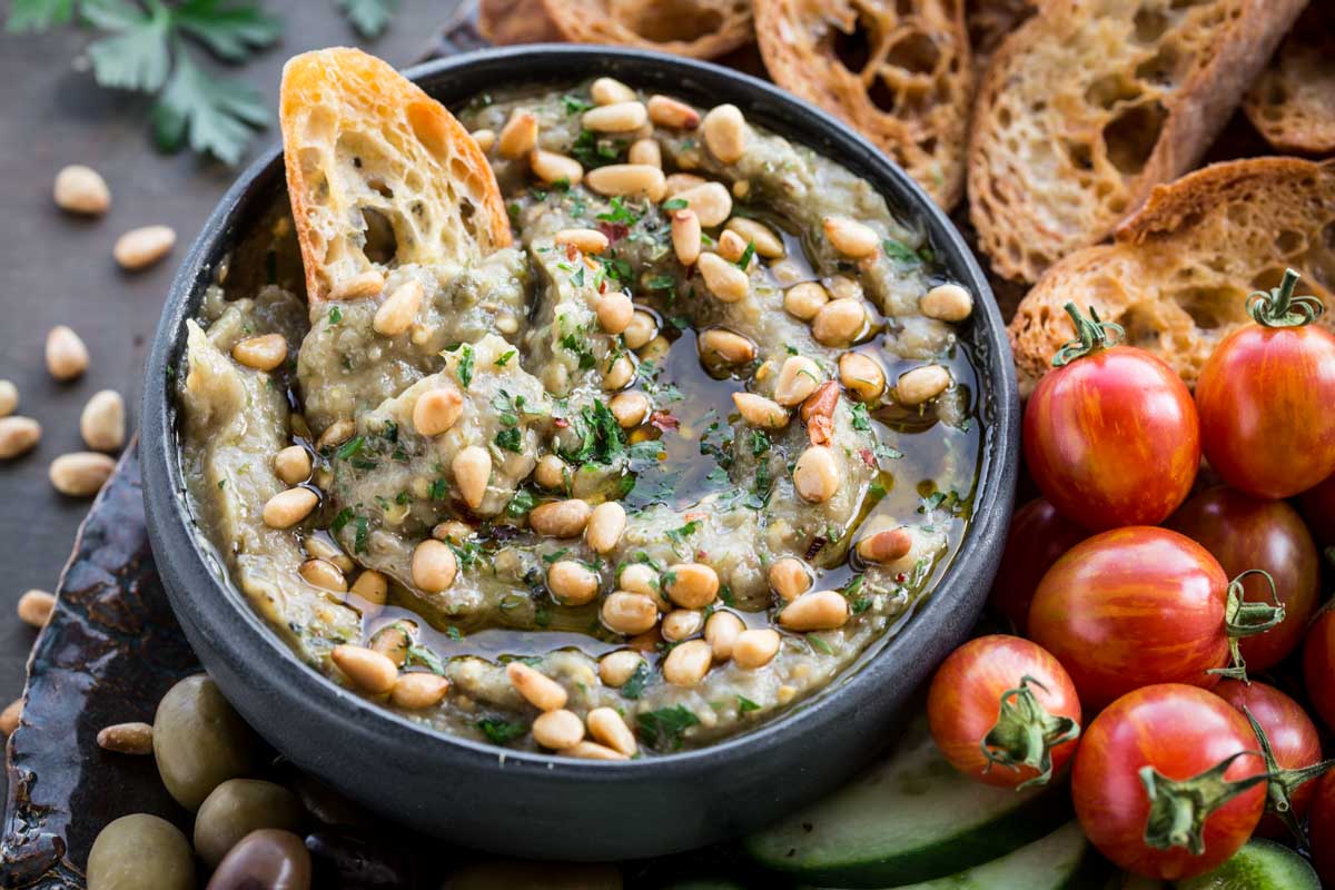 Dipping bread into eggplant dip - A piece of toasted bread dipped into a bowl of creamy smoky eggplant dip topped with pine nuts, with fresh tomatoes and cucumbers in the background.