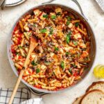 Pasta coated in bright red tomato sauce with golden roasted eggplant, melted mozzarella cubes, and fresh basil, served in a gray skillet with a wooden spoon, surrounded by fresh bread, basil leaves, and a block of cheese.