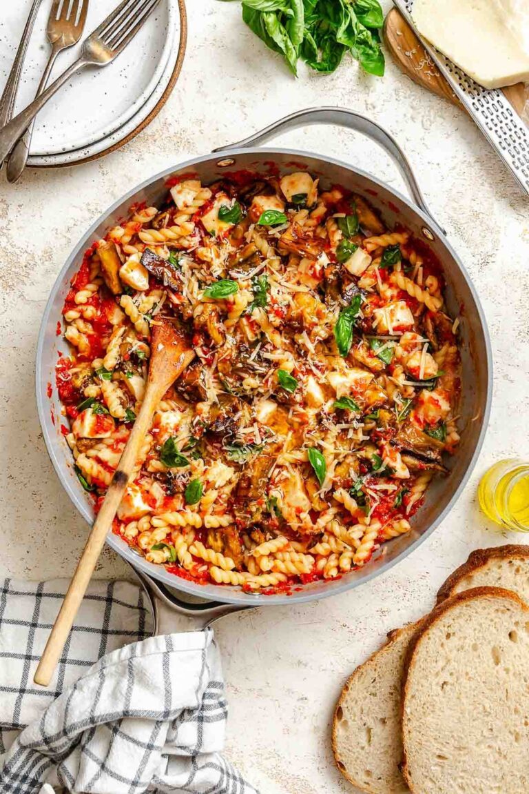 Pasta coated in bright red tomato sauce with golden roasted eggplant, melted mozzarella cubes, and fresh basil, served in a gray skillet with a wooden spoon, surrounded by fresh bread, basil leaves, and a block of cheese.
