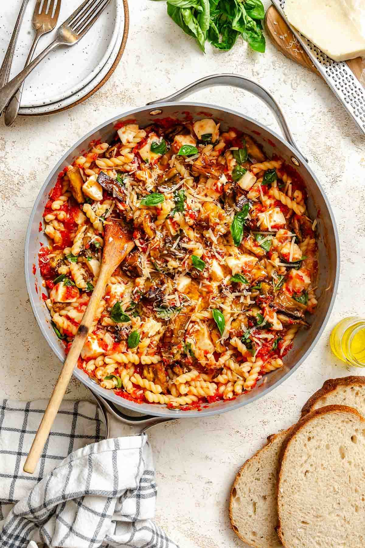 Pasta coated in bright red tomato sauce with golden roasted eggplant, melted mozzarella cubes, and fresh basil, served in a gray skillet with a wooden spoon, surrounded by fresh bread, basil leaves, and a block of cheese.