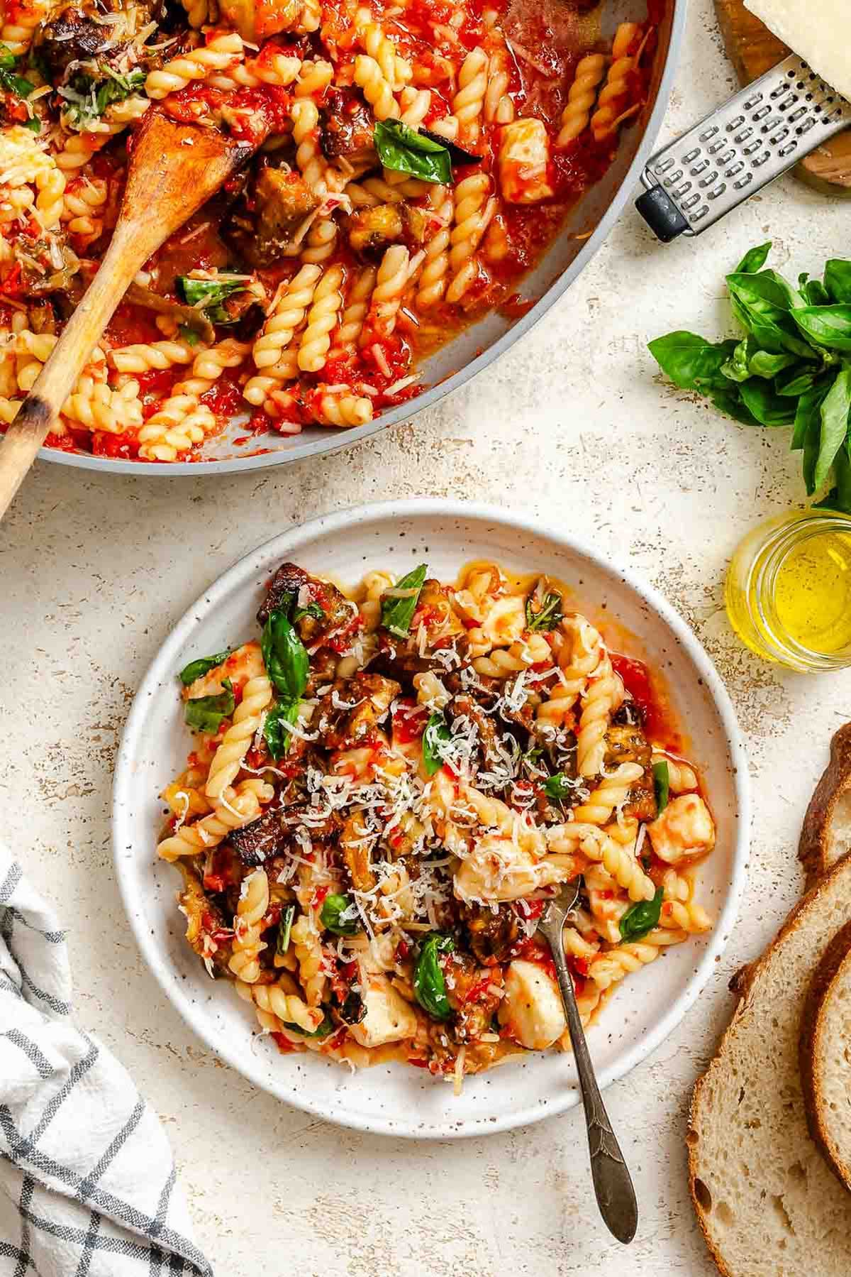 White ceramic plate of pasta with roasted eggplant, mozzarella, basil, and Parmesan, next to the skillet of sauce and fresh bread slices, garnished with fresh herbs.