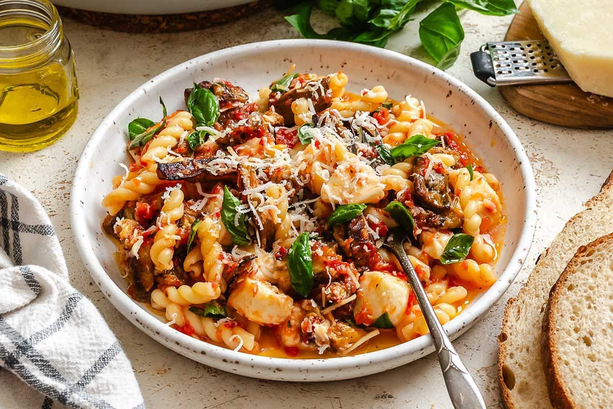 White speckled bowl filled with pasta, roasted eggplant, melted mozzarella, and fresh basil, topped with a sprinkle of grated Parmesan, with bread slices and olive oil jar nearby.