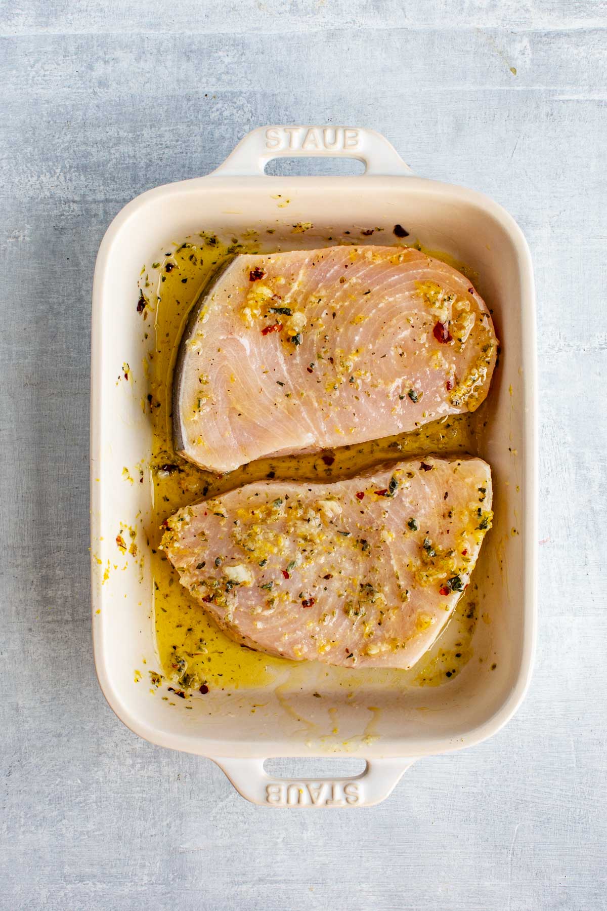 Two swordfish steaks marinating in olive oil, lemon zest, garlic, and herbs inside a cream-colored Staub baking dish.