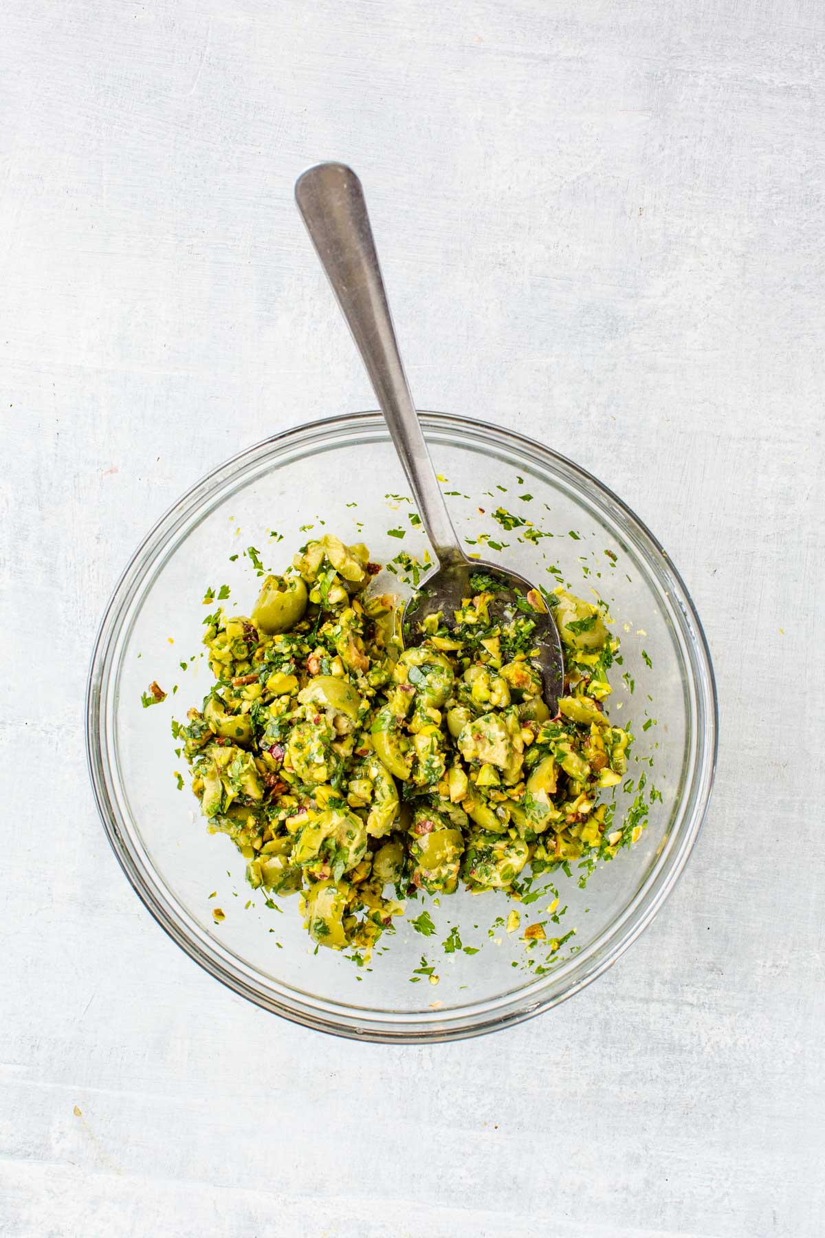A glass bowl filled with a chunky mixture of Castelvetrano olives, pistachios, parsley, garlic, and lemon with a spoon for serving.