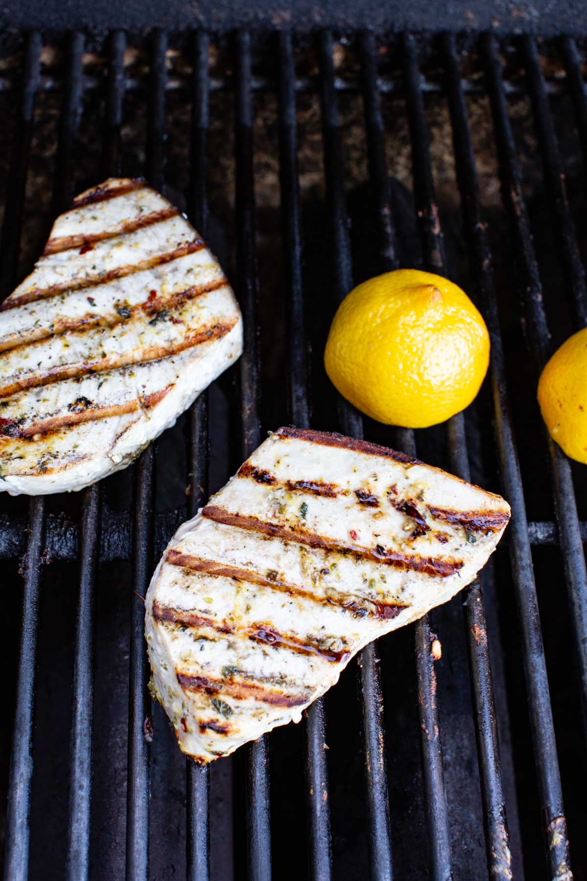 Swordfish steaks cooking on hot grill grates next to fresh lemons, showing golden brown sear marks.
