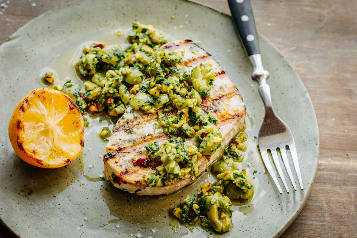 A swordfish steak topped generously with olive pistachio compote, juices pooling around, with a grilled lemon on the side.