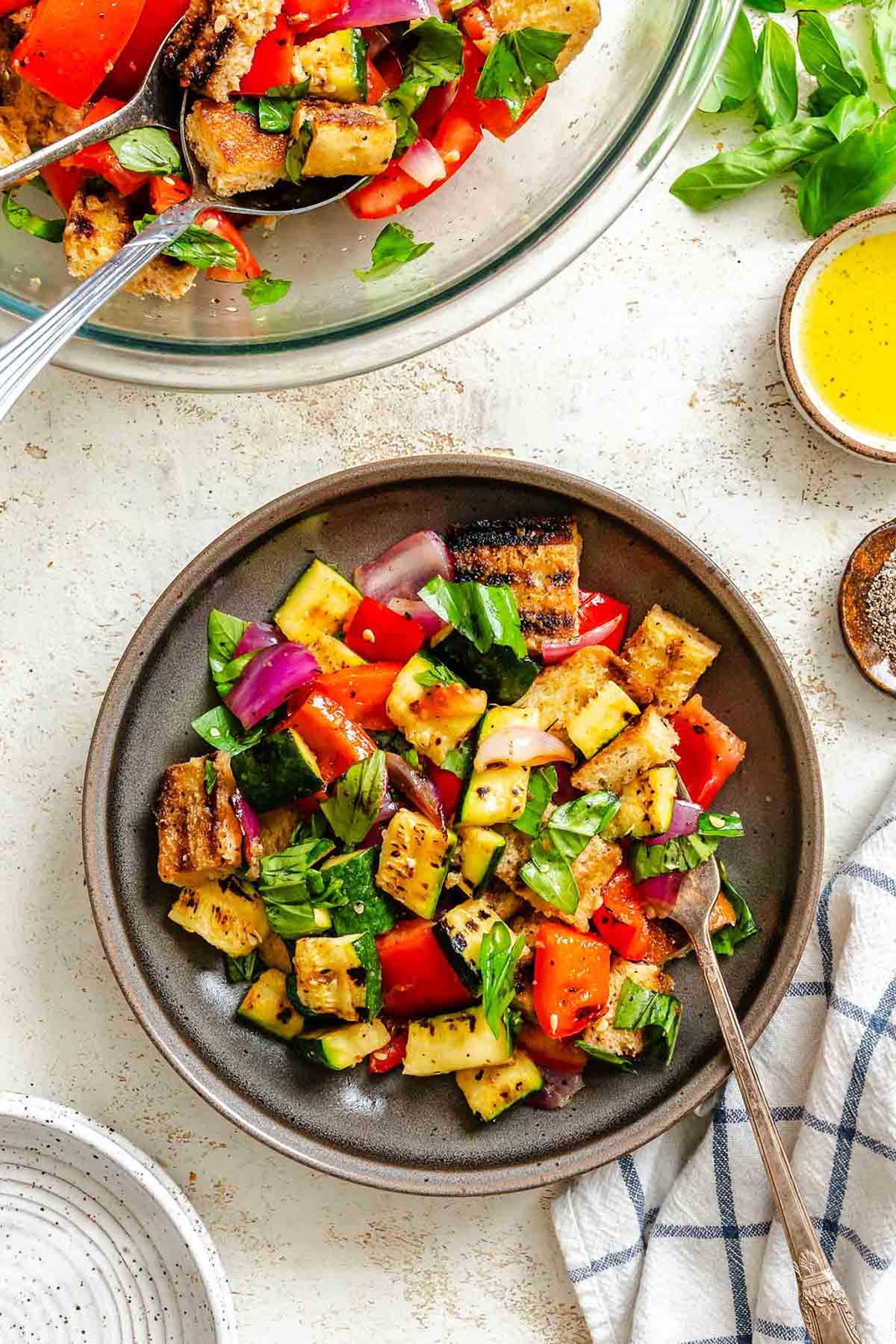 Fresh grilled panzanella salad with tomatoes, zucchini, red onion, and charred bread cubes in a mixing bowl and a serving plate with basil and vinaigrette on the side.