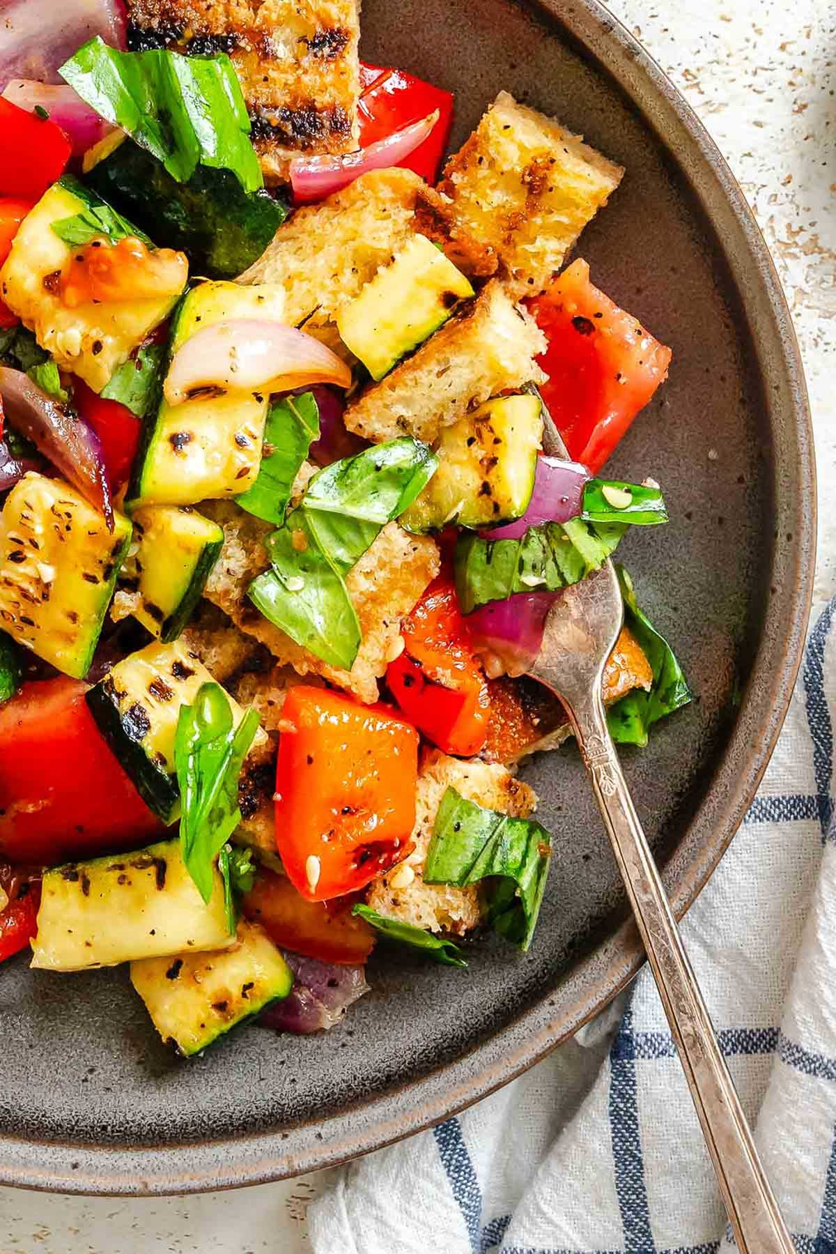 Close-up of grilled panzanella on plate - Crisp grilled bread cubes, zucchini, bell peppers, red onion, and torn basil in a rustic plate with a vintage fork.