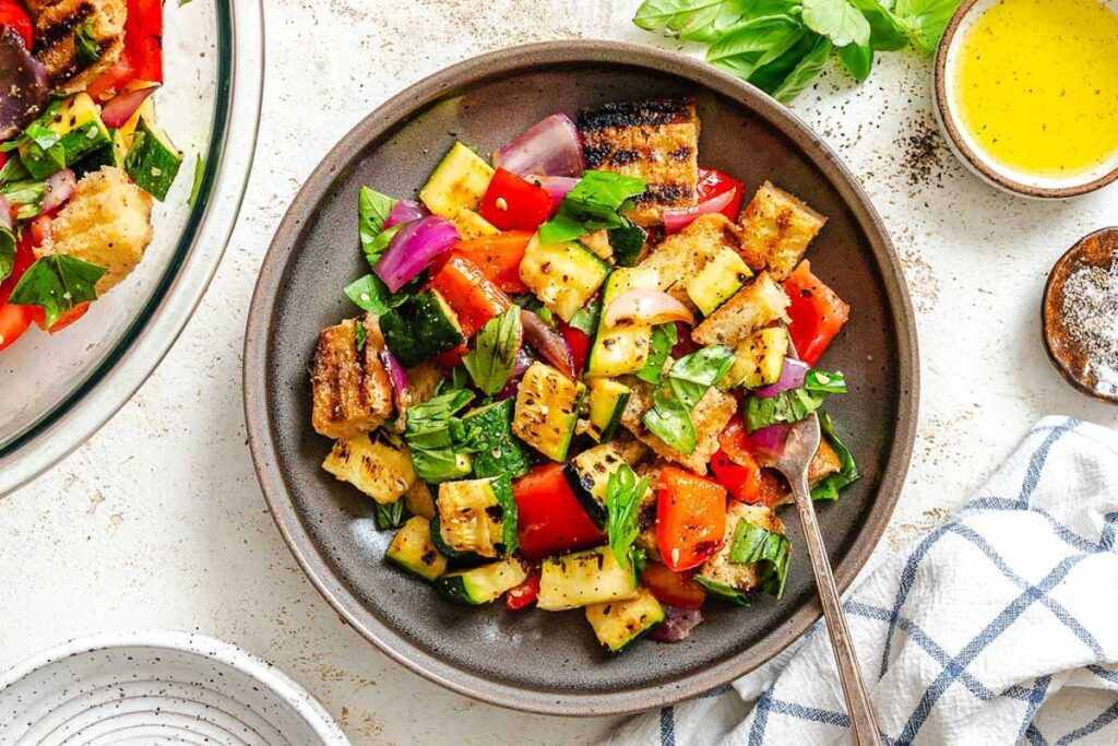 Overhead shot of plated salad and dressing - Top-down view of colorful grilled vegetable and bread salad in a dark plate, surrounded by fresh basil, pepper, and dressing.