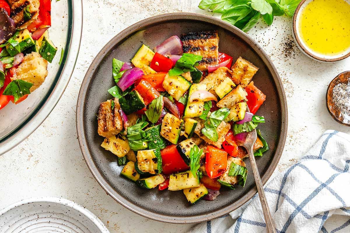 Overhead shot of plated salad and dressing – Top-down view of colorful grilled vegetable and bread salad in a dark plate, surrounded by fresh basil, pepper, and dressing.