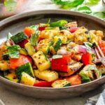 Side view of grilled panzanella - Bright plate of grilled panzanella salad with chunky vegetables and herbs, next to a bowl of dressing and bunch of fresh basil.