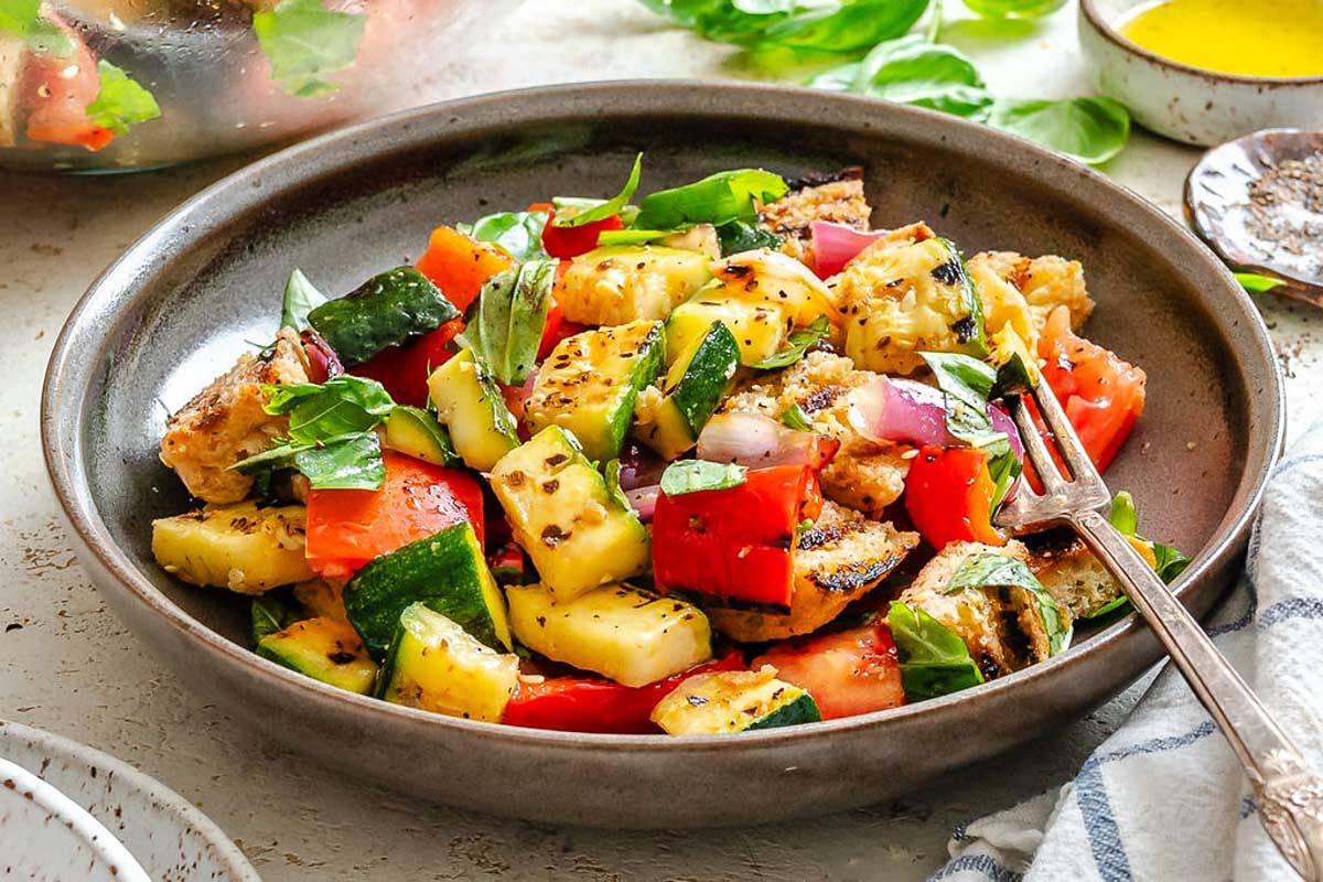 Side view of grilled panzanella - Bright plate of grilled panzanella salad with chunky vegetables and herbs, next to a bowl of dressing and bunch of fresh basil.