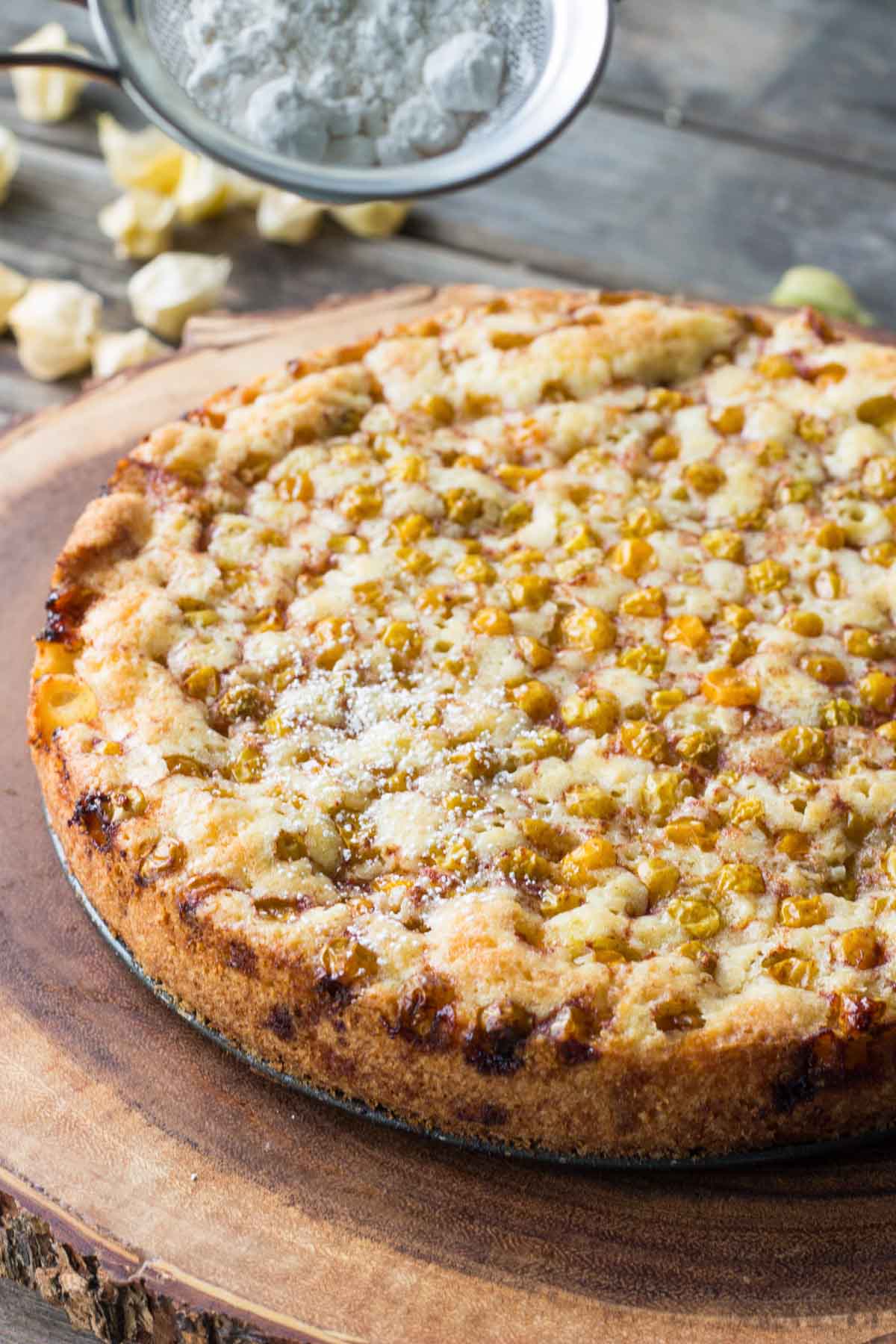 A freshly baked ground cherry cake on a wooden board, topped with golden fruit and being dusted with powdered sugar through a sifter.