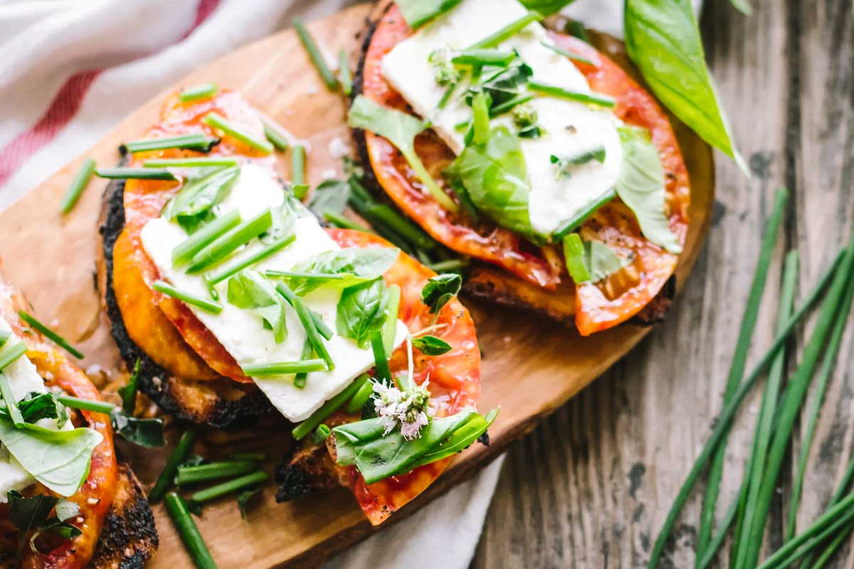 Overhead view of heirloom tomato toasts on a rustic wooden surface, each slice garnished with feta, basil, and chopped chives, with scattered herbs and a striped dish towel nearby.
