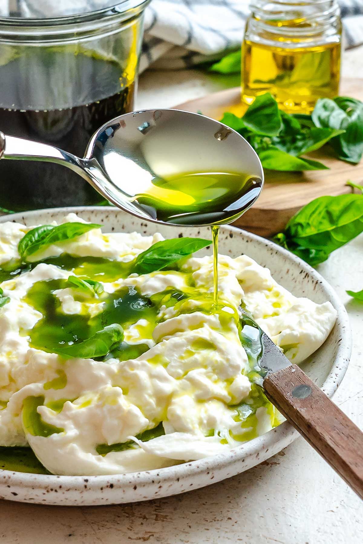 Spoon drizzling basil oil over burrata and basil leaves on a white plate.
