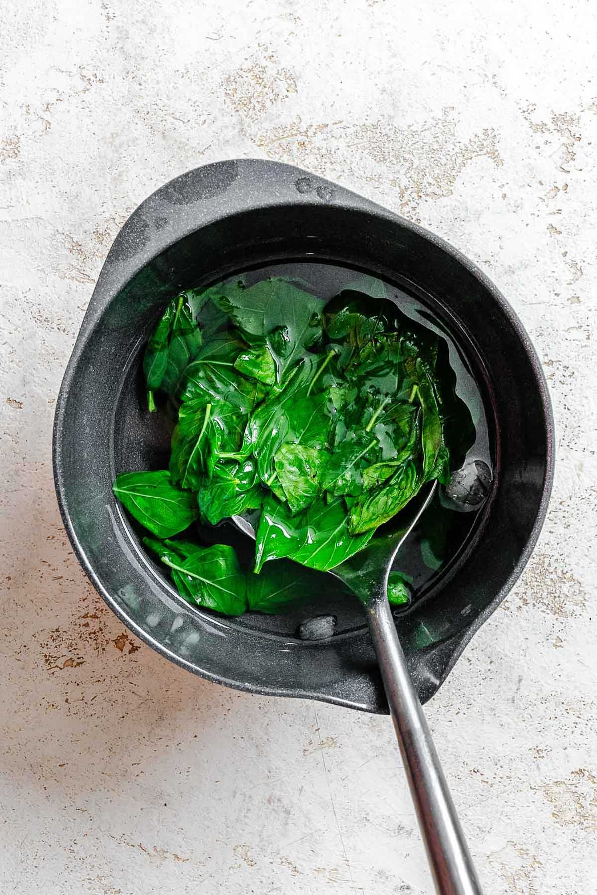 Cooling blanched basil in a black bowl of ice water, with a slotted spoon lifting the vibrant leaves.
