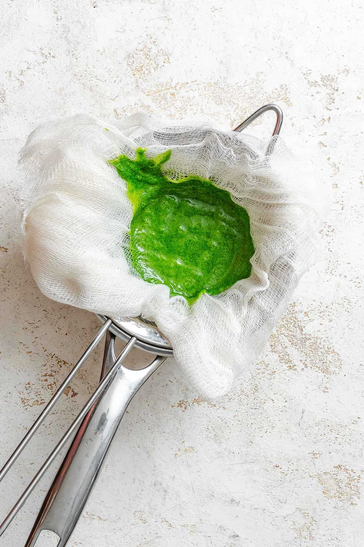 Straining basil puree through layered cheesecloth over a fine sieve to remove solids.