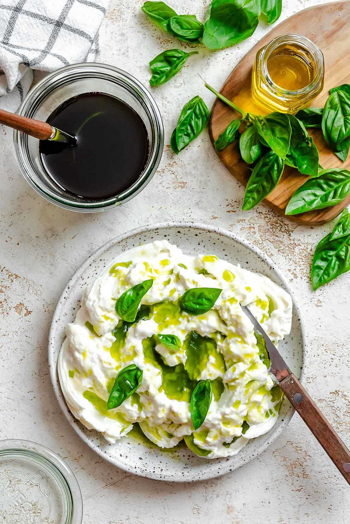 Burrata with basil oil drizzle, served on a speckled plate with fresh basil leaves on top.