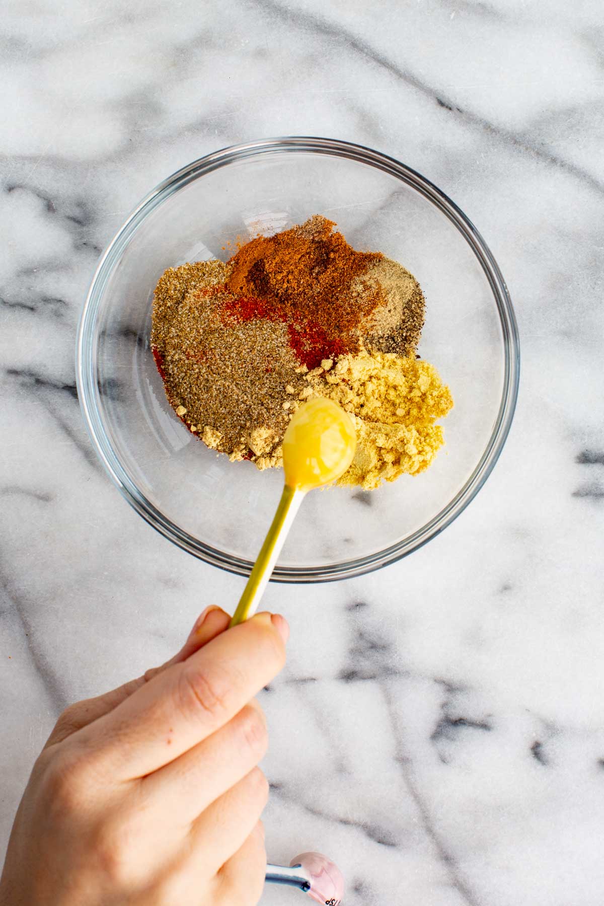 Hand holding a yellow measuring spoon above a glass bowl filled with celery salt, mustard powder, paprika, and other spices for homemade seasoning.