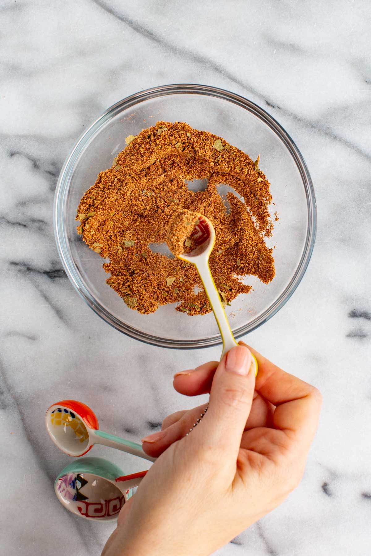 Hand holding a spoonful of freshly mixed Old Bay-style seasoning in a glass bowl, with visible flakes of bay leaf and a reddish-brown color.