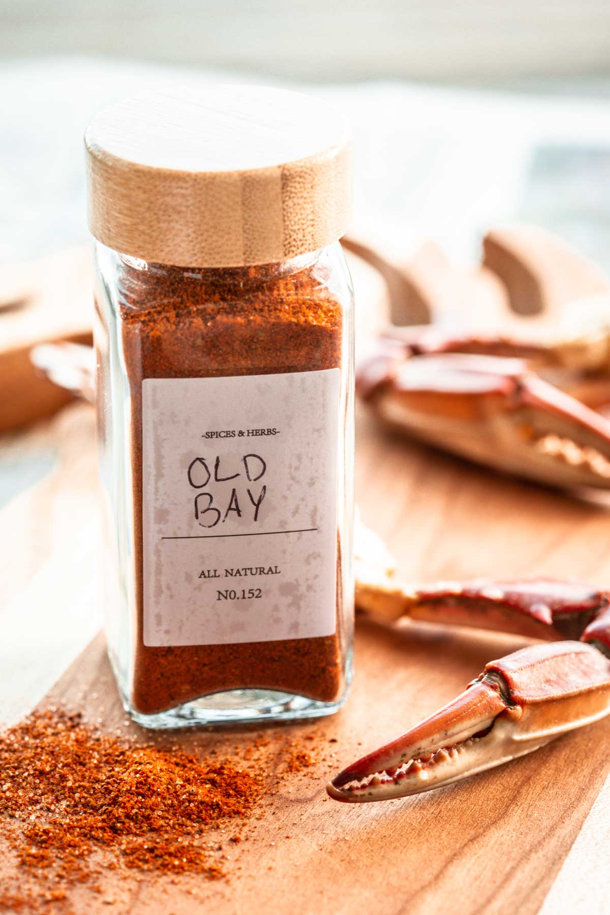 Homemade Old Bay seasoning in a spice jar with bamboo lid on a wooden board, surrounded by steamed crab claws and a dusting of spice.