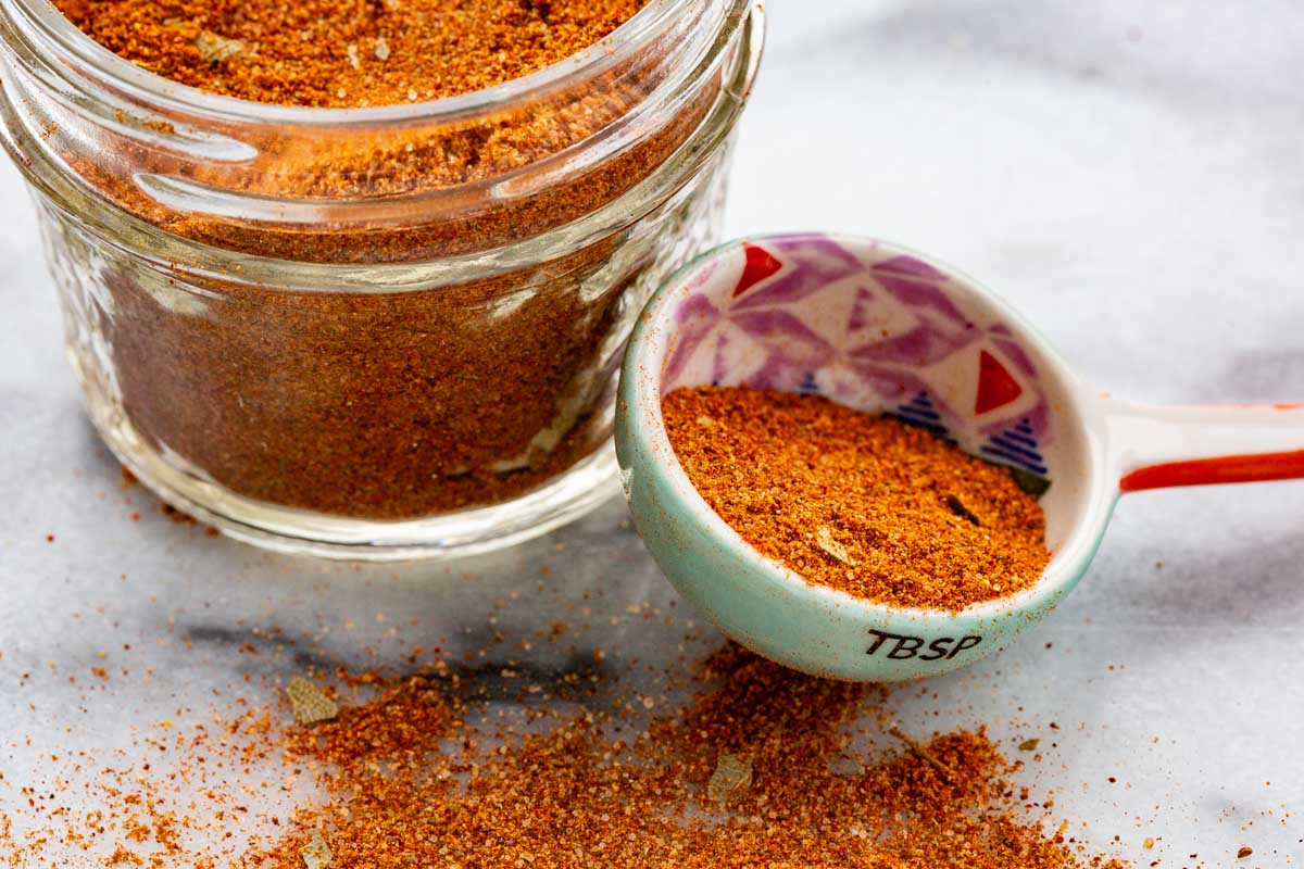 Close-up of a colorful ceramic tablespoon filled with homemade seasoning next to a small glass jar on a marble surface.