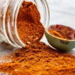 Homemade Old Bay seasoning spilling out of a tipped-over glass jar onto a white marble counter, showing fine texture and bay leaf flakes.