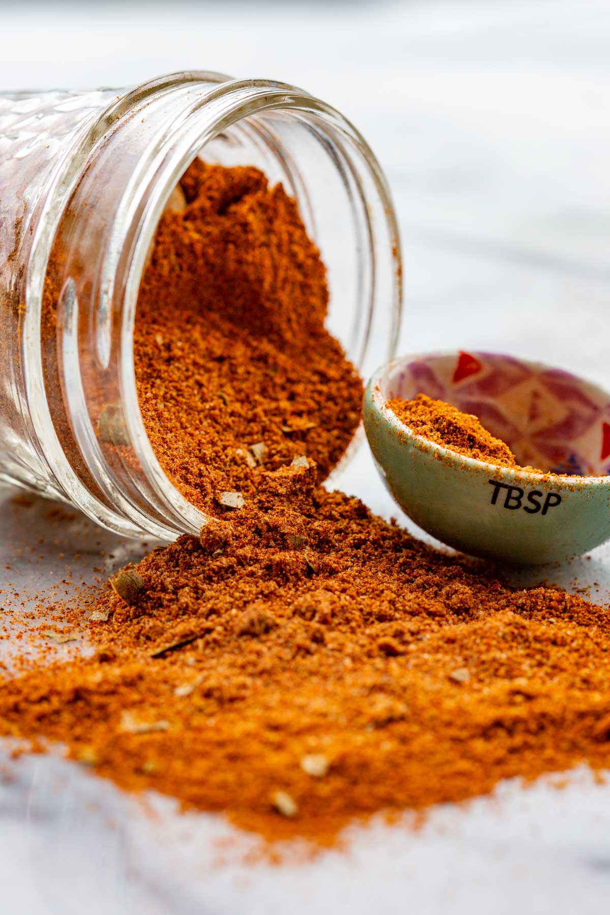 Homemade Old Bay seasoning spilling out of a tipped-over glass jar onto a white marble counter, showing fine texture and bay leaf flakes.