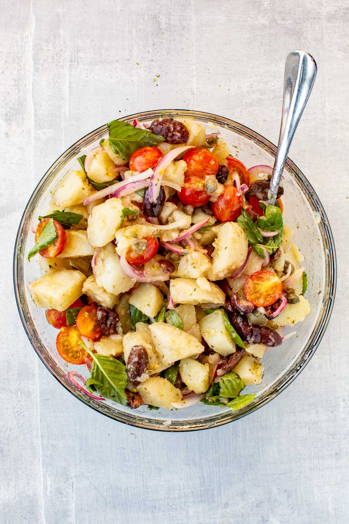 Glass bowl with insalata pantesca – Chunks of boiled potatoes mixed with cherry tomatoes, black olives, capers, onion slices, and fresh basil leaves, lightly dressed with olive oil.