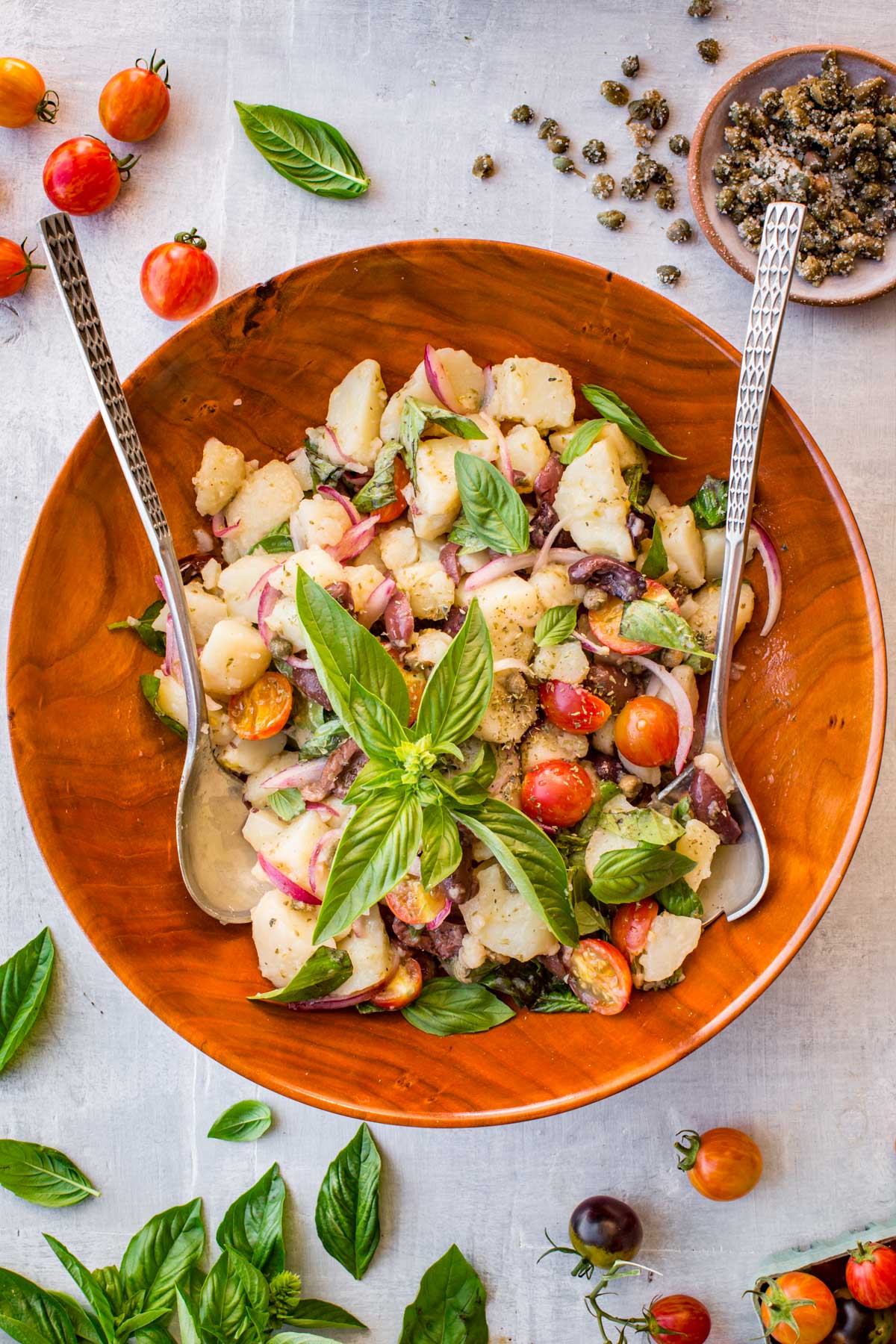 Wooden bowl of Sicilian potato salad - Insalata pantesca garnished with fresh basil sprigs, surrounded by cherry tomatoes and a small dish of salt-packed capers.