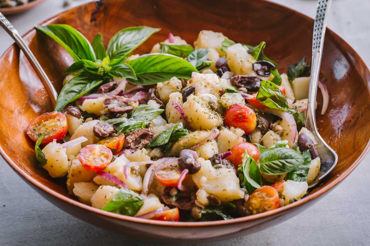 Wide bowl of insalata pantesca - A large serving of Sicilian potato salad in a wooden bowl, with bright basil leaves and glossy cherry tomatoes.