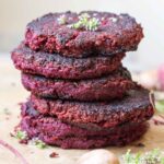 Stack of beet and lentil burgers - Four deep red lentil and beet patties stacked high on parchment paper, garnished with fresh herbs and surrounded by mushrooms and beet stems.