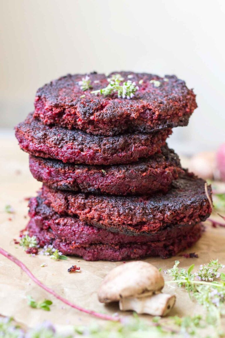 Stack of beet and lentil burgers – Four deep red lentil and beet patties stacked high on parchment paper, garnished with fresh herbs and surrounded by mushrooms and beet stems.