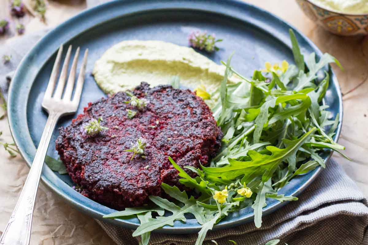 Beet burger plated with arugula and sauce - Seared beet and lentil burger on a blue plate with fresh arugula and a creamy green sauce, garnished with edible flowers.