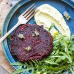 Overhead of beet burger meal - Top-down view of a single beet burger with whipped green sauce and arugula on a blue plate, surrounded by herbs and napkin.