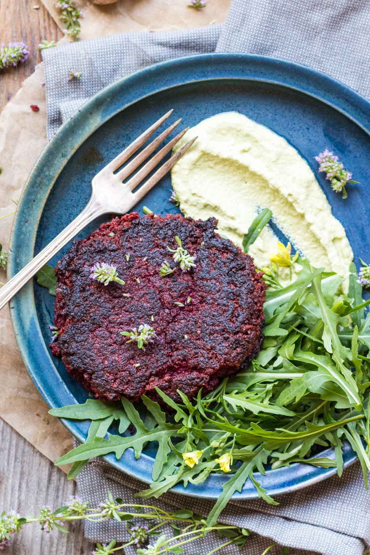 Overhead of beet burger meal - Top-down view of a single beet burger with whipped green sauce and arugula on a blue plate, surrounded by herbs and napkin.