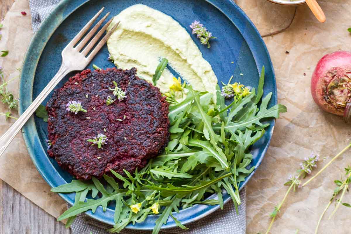 Wide shot of plated beet burger – Lentil and beet burger served with arugula and sauce on a rustic plate, styled with fresh herbs, parchment, and a raw beet.