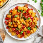 Overhead view of a plate of Pasta Ponza with rigatoni, tomatoes, fresh basil, and capers, next to fresh basil leaves.