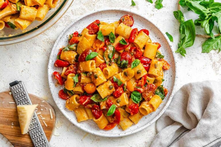 Overhead view of a plate of Pasta Ponza with rigatoni, tomatoes, fresh basil, and capers, next to fresh basil leaves.