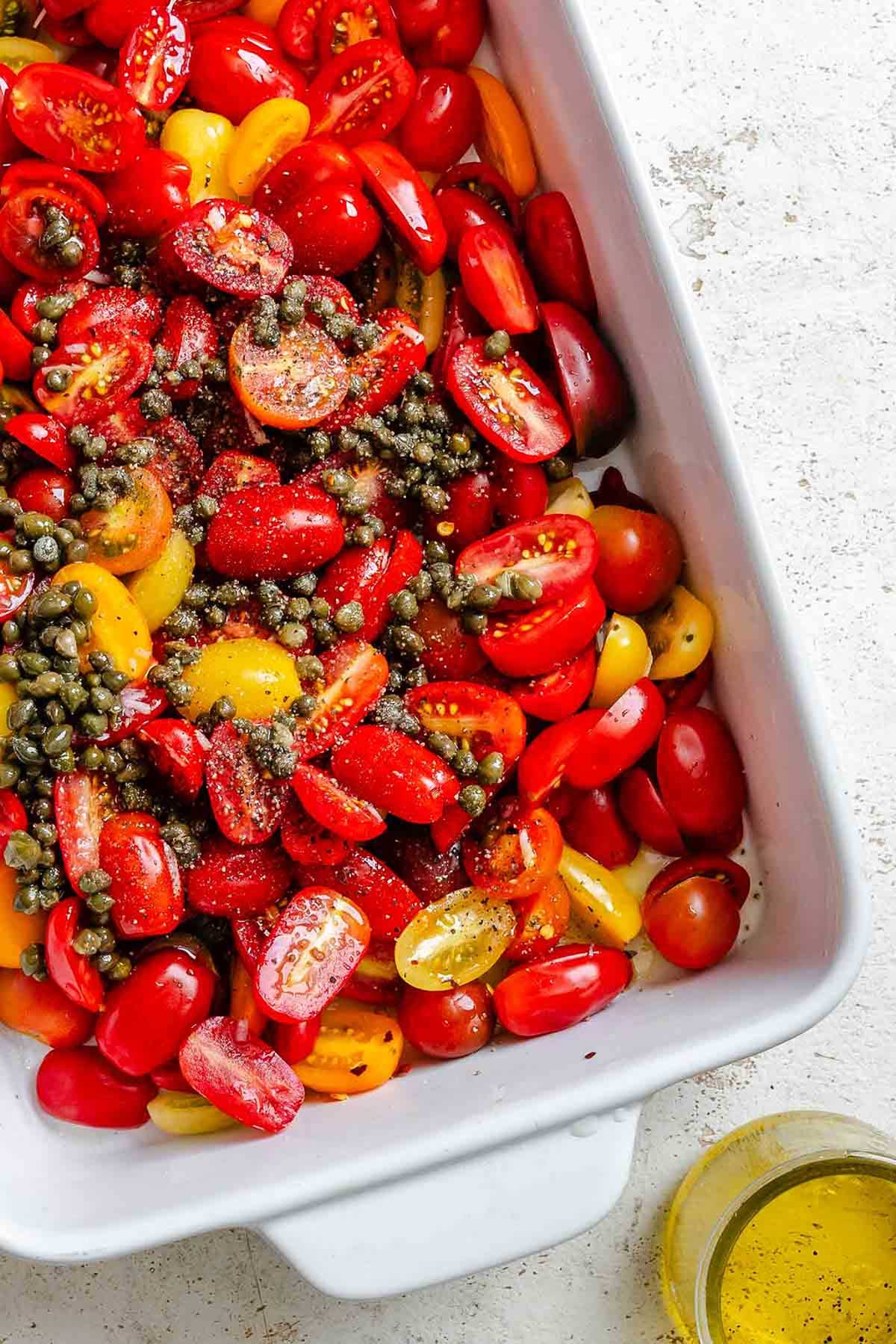 Halved cherry tomatoes and yellow grape tomatoes in a white baking dish, tossed with capers, olive oil, salt, and pepper.