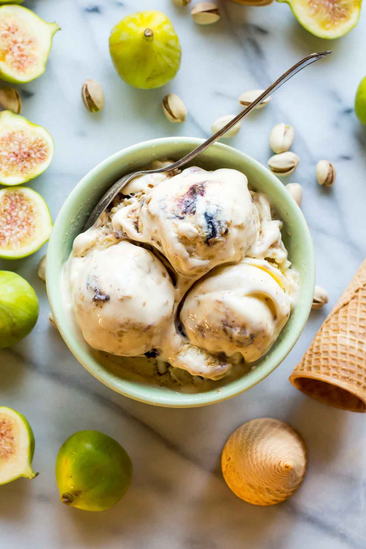 A green bowl filled with three scoops of ricotta gelato swirled with roasted figs and pistachio, with a spoon tucked in. Fresh figs, pistachios, and an ice cream cone are scattered on the marble surface.