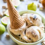 A bowl of ricotta, pistachio and fig gelato with an upside-down waffle cone placed on top, surrounded by halved figs and pistachios.
