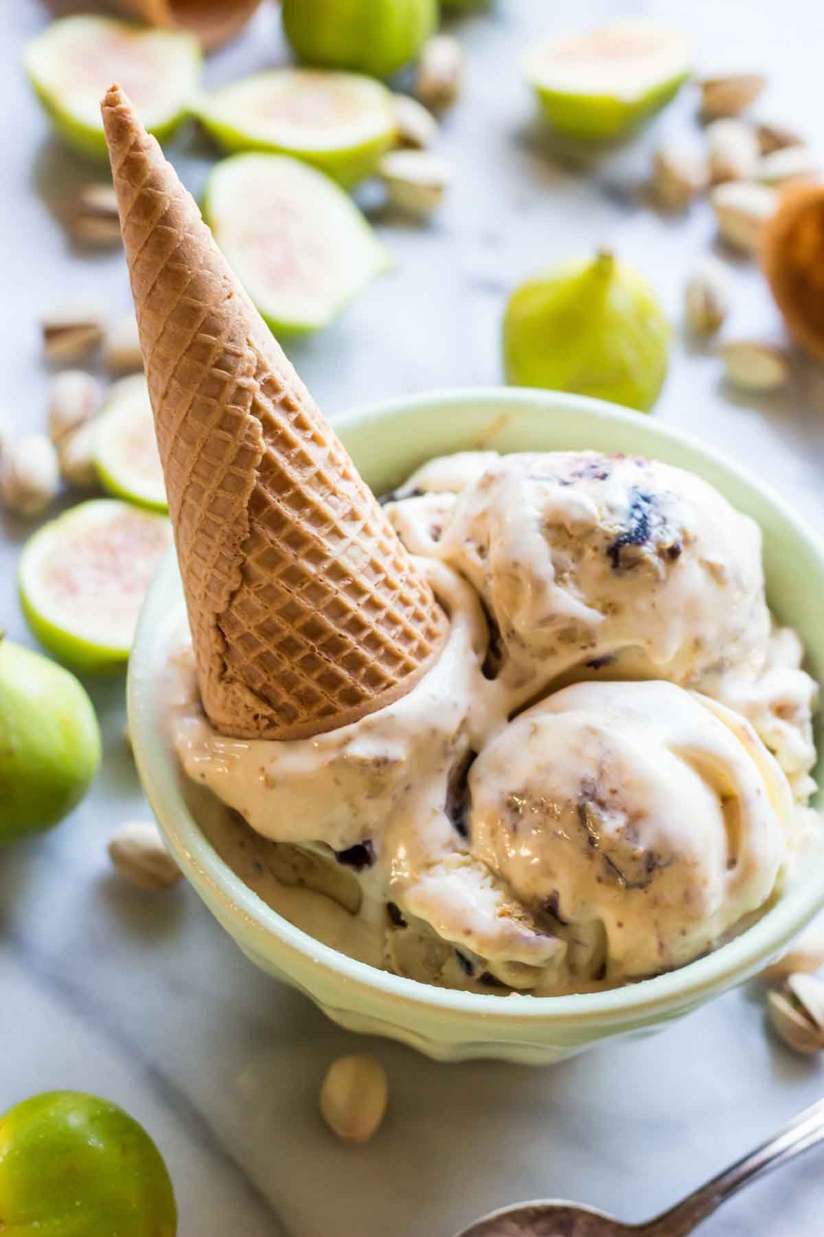 A bowl of ricotta, pistachio and fig gelato with an upside-down waffle cone placed on top, surrounded by halved figs and pistachios.