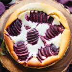 Overhead view of a whole Italian ricotta plum cheesecake on a wooden board, with fanned plum slices arranged in a starburst pattern over creamy filling.