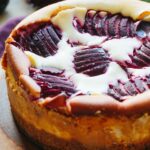 Close-up of a baked Italian ricotta plum cheesecake on a wooden board, showing the golden crust and fanned dark plum slices on top with creamy filling peeking through.