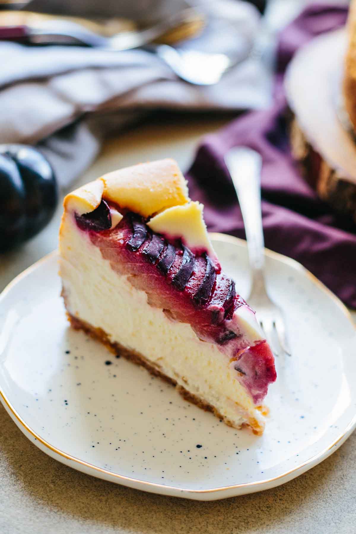 Single slice of Italian ricotta plum cheesecake on a white speckled plate, showing creamy layers, graham cracker crust, and juicy baked plum topping.