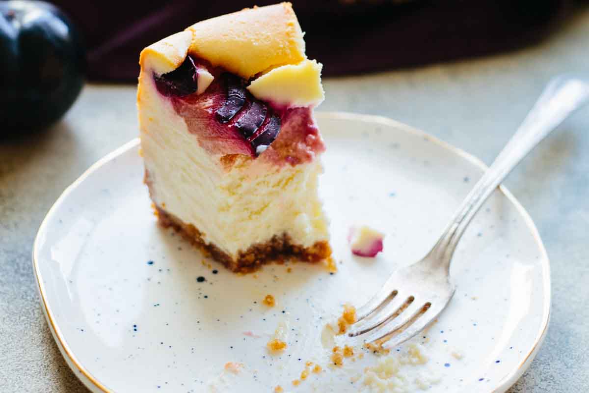 Partially eaten slice of Italian ricotta plum cheesecake on a white speckled plate with a fork, showing the rich, creamy texture and cinnamon-dusted plum topping.